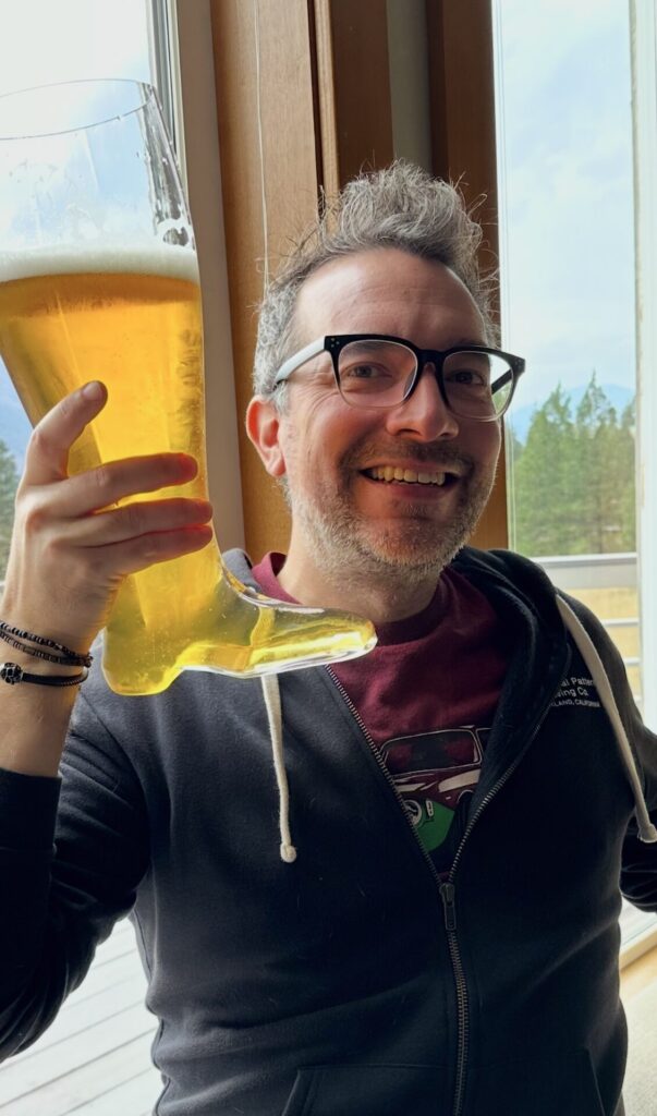 A fair skinned man with dark eyes, holding a Das Boot full of golden beer. The man is wearing dark rimmed glasses and a broad smile